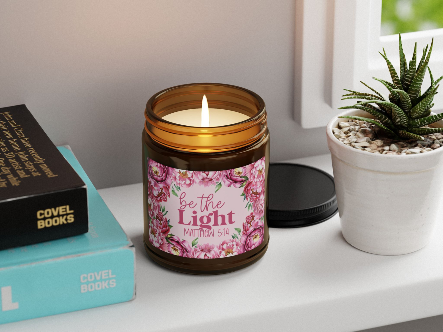 Lit candle in a glass jar with floral label reading, "Be the Light, Matthew 5:14." Beside it are books and a potted succulent on a white shelf.