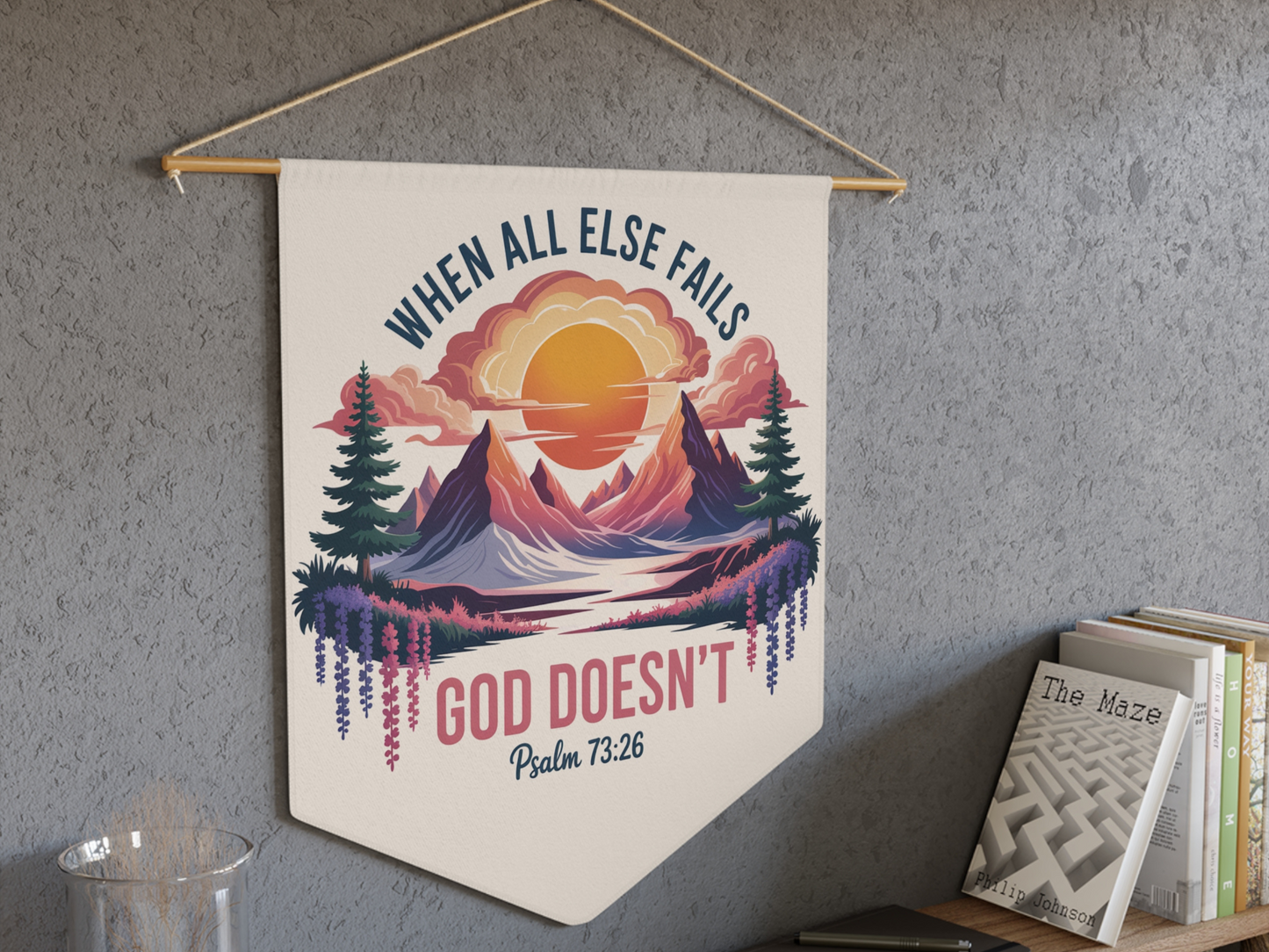 Wall art hanging with a vibrant landscape of mountains and sunset reads, "When all else fails God doesn't" with Psalm 73:26. Books are on a nearby shelf.