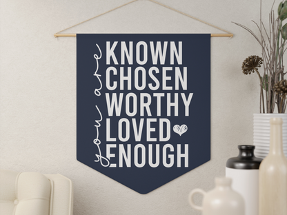 Wall banner with motivational text: "You are known, chosen, worthy, loved, enough" in bold white letters on navy. Cozy room with a plant and vases adds warmth.