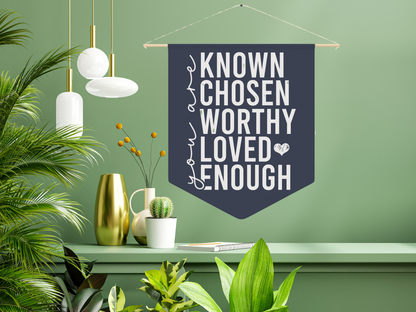 A stylish room with a green wall, plants, and modern white and gold pendant lights. A dark banner reads, "You are known, chosen, worthy, loved, enough."