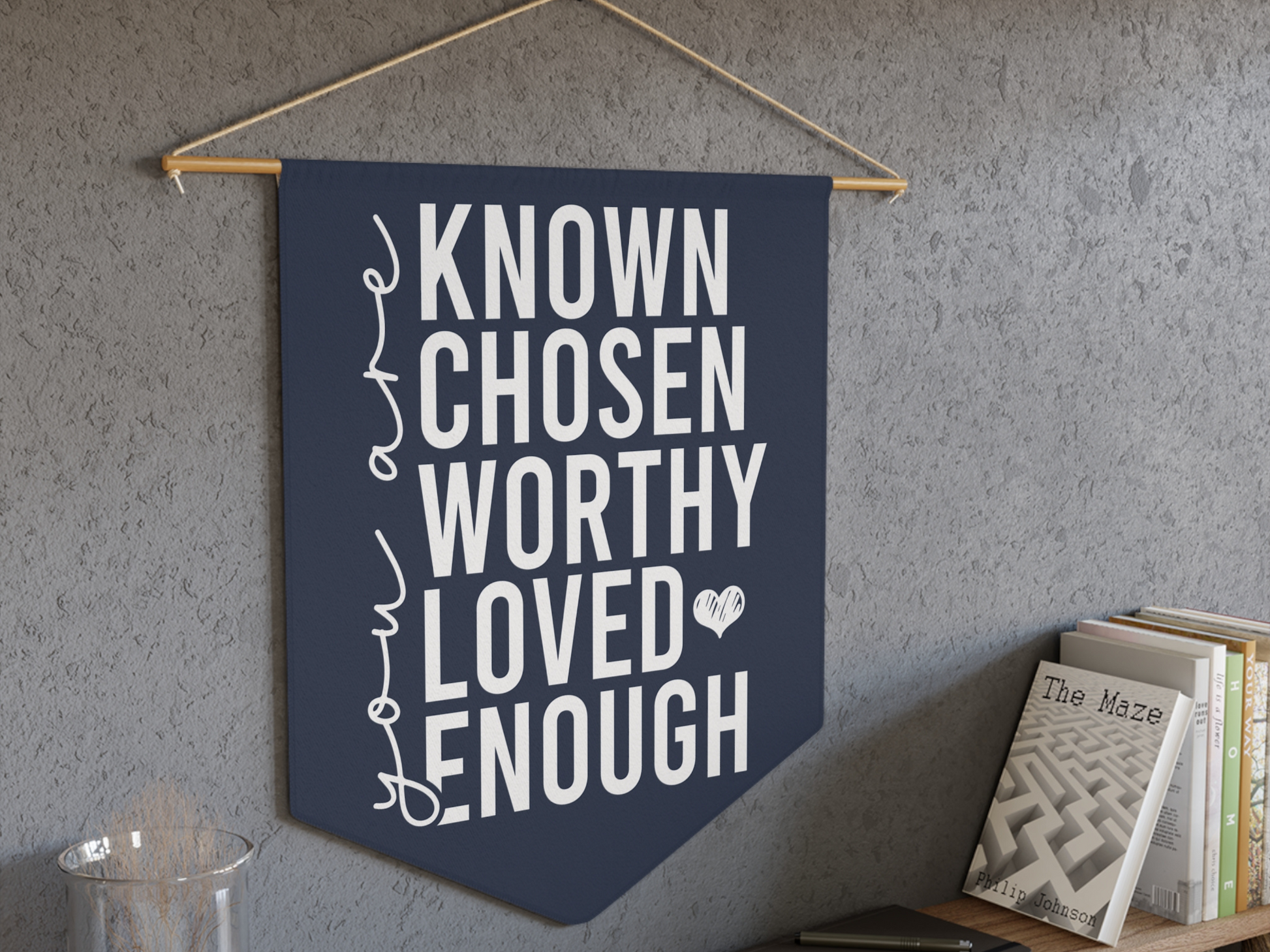 A navy wall banner with motivational words: "KNOWN, CHOSEN, WORTHY, LOVED, ENOUGH" in white text and a small heart. A shelf with books is beside it.