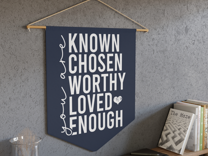 A navy wall banner with motivational words: "KNOWN, CHOSEN, WORTHY, LOVED, ENOUGH" in white text and a small heart. A shelf with books is beside it.