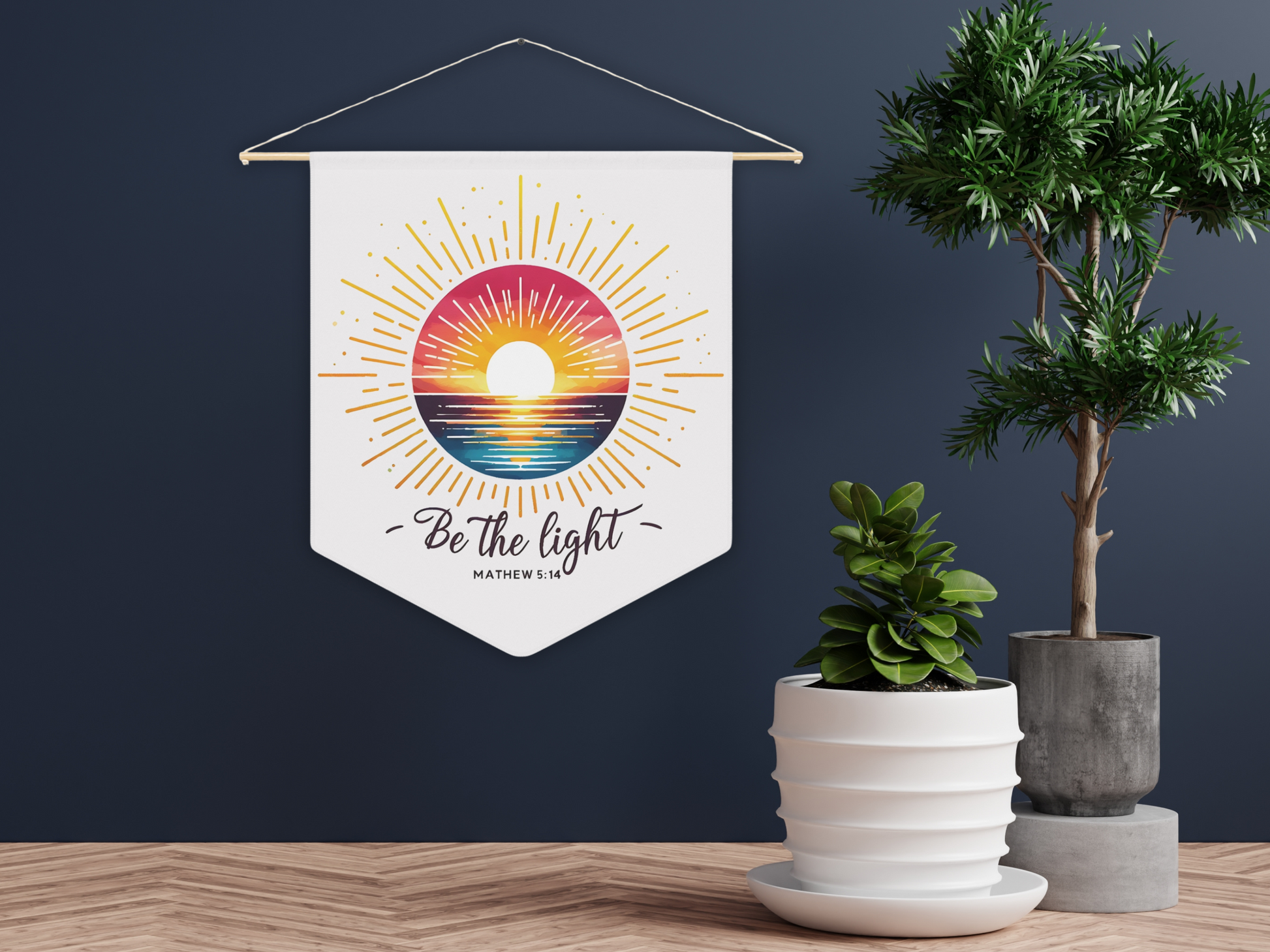 A wall hanging with a colorful sunrise over water, text "Be the light, Matthew 5:14" in elegant script. Nearby are potted plants on a wooden floor.
