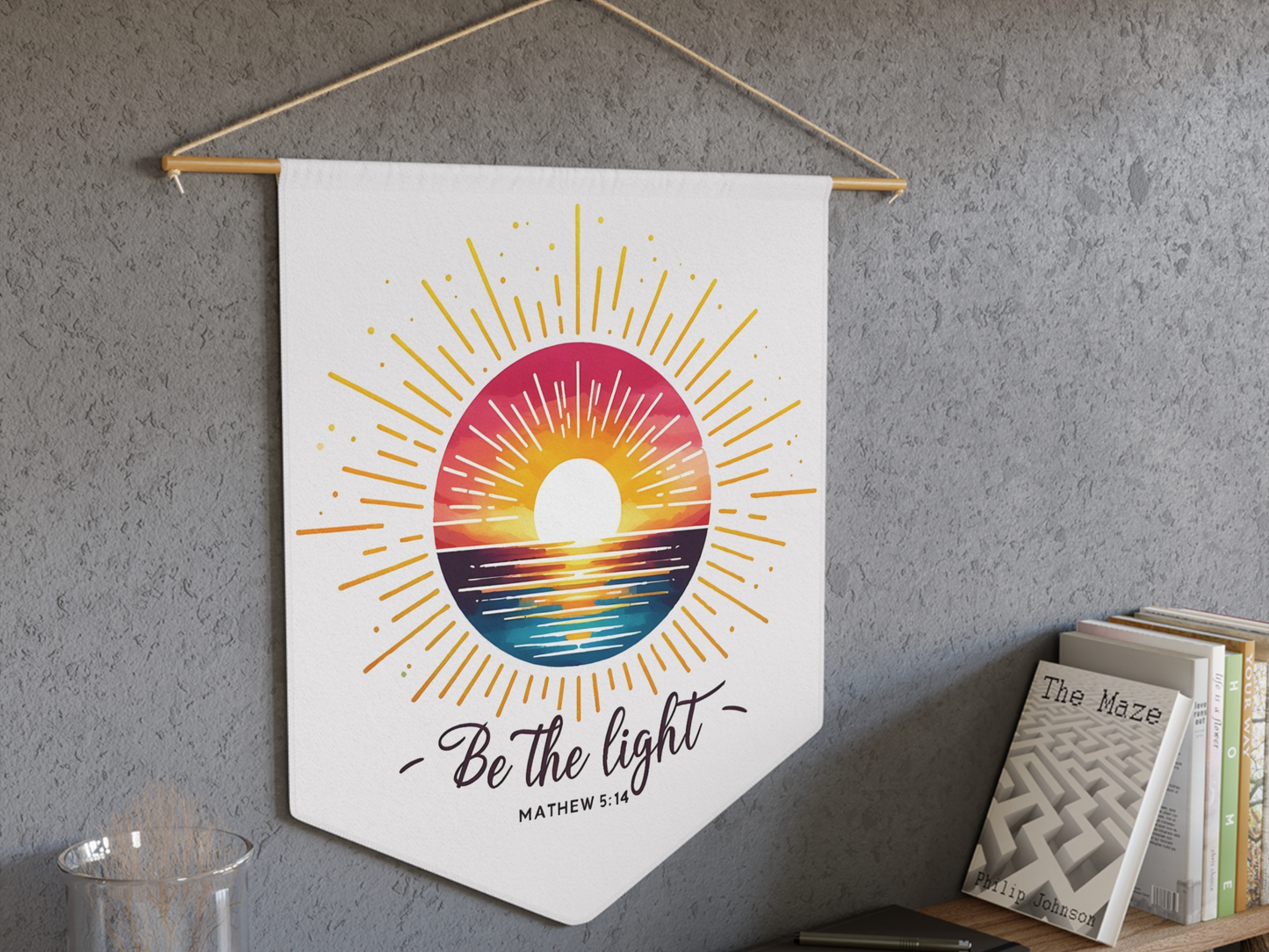 Wall hanging with a vibrant sunburst rising over waves, captioned "Be the light". Set against a textured gray wall above a bookshelf.