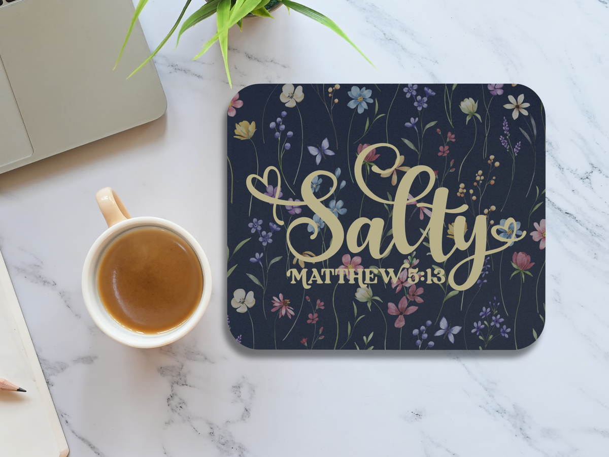 Mouse pad with a floral design and the text "Salty Matthew 5:13" in elegant script. It's placed on a marble table beside a cup of coffee.