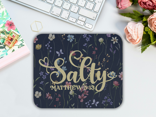 A floral mousepad on a desk features "Salty Matthew 5:13" in elegant script. Nearby is a keyboard, a floral notepad, and pink roses, creating a fresh, creative vibe.