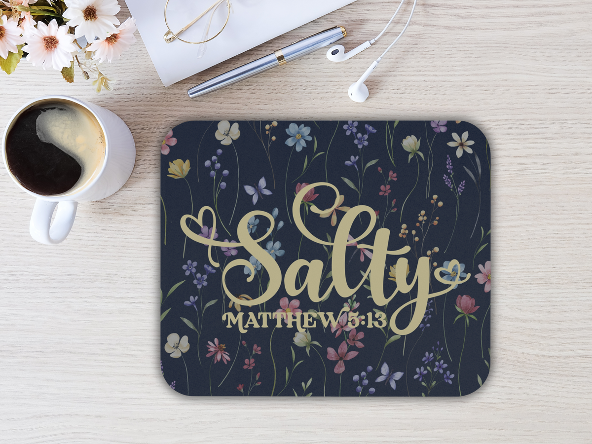 A mousepad with "Salty" and "Matthew 5:13" script on a dark, floral background. Nearby are flowers, glasses, a pen, earbuds, and a coffee mug. Sophisticated and calming ambiance.