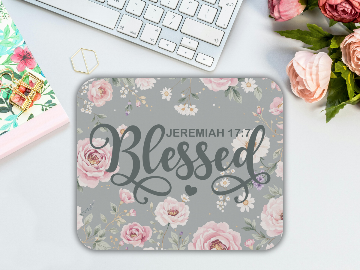 A floral-themed mouse pad with the word "Blessed" and "Jeremiah 17:7" on a gray background, surrounded by pink flowers and greenery. A keyboard and roses are nearby, creating a serene workspace vibe.