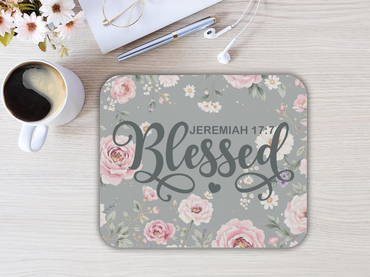 A floral mouse pad with pink roses and white daisies features the text "Jeremiah 17:7 Blessed" in elegant script. Nearby, coffee, flowers, and earbuds rest on a light wood table, creating a calm and inspirational tone.