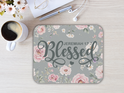 A floral mouse pad with pink roses and white daisies features the text "Jeremiah 17:7 Blessed" in elegant script. Nearby, coffee, flowers, and earbuds rest on a light wood table, creating a calm and inspirational tone.
