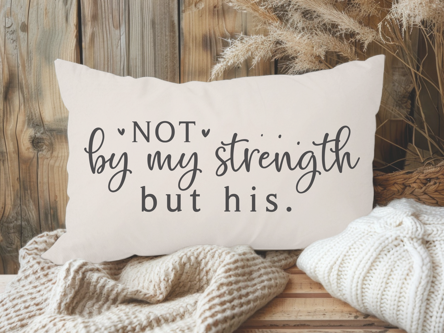 A decorative pillow with the words "Not by my strength but his" in elegant script lies on a wooden surface, surrounded by cozy blankets and dried plants, creating a warm, rustic ambiance.