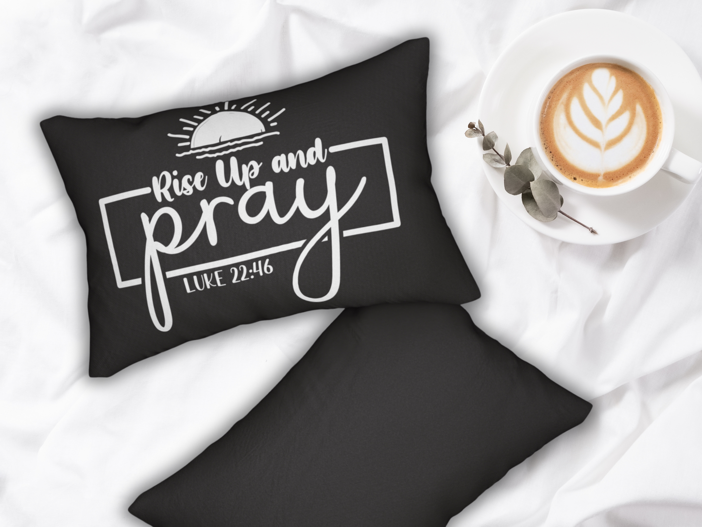 A black pillow with "Rise Up and Pray, Luke 22:46" in white text next to a latte with leaf art and eucalyptus on a white bed. Calm and reflective tone.