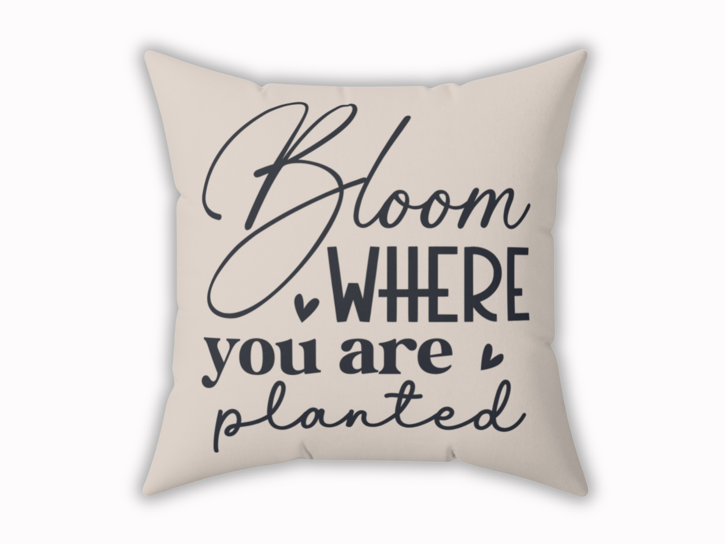 A beige throw pillow features the motivational phrase "Bloom where you are planted" in elegant, navy script. Heart accents add a warm, uplifting tone.