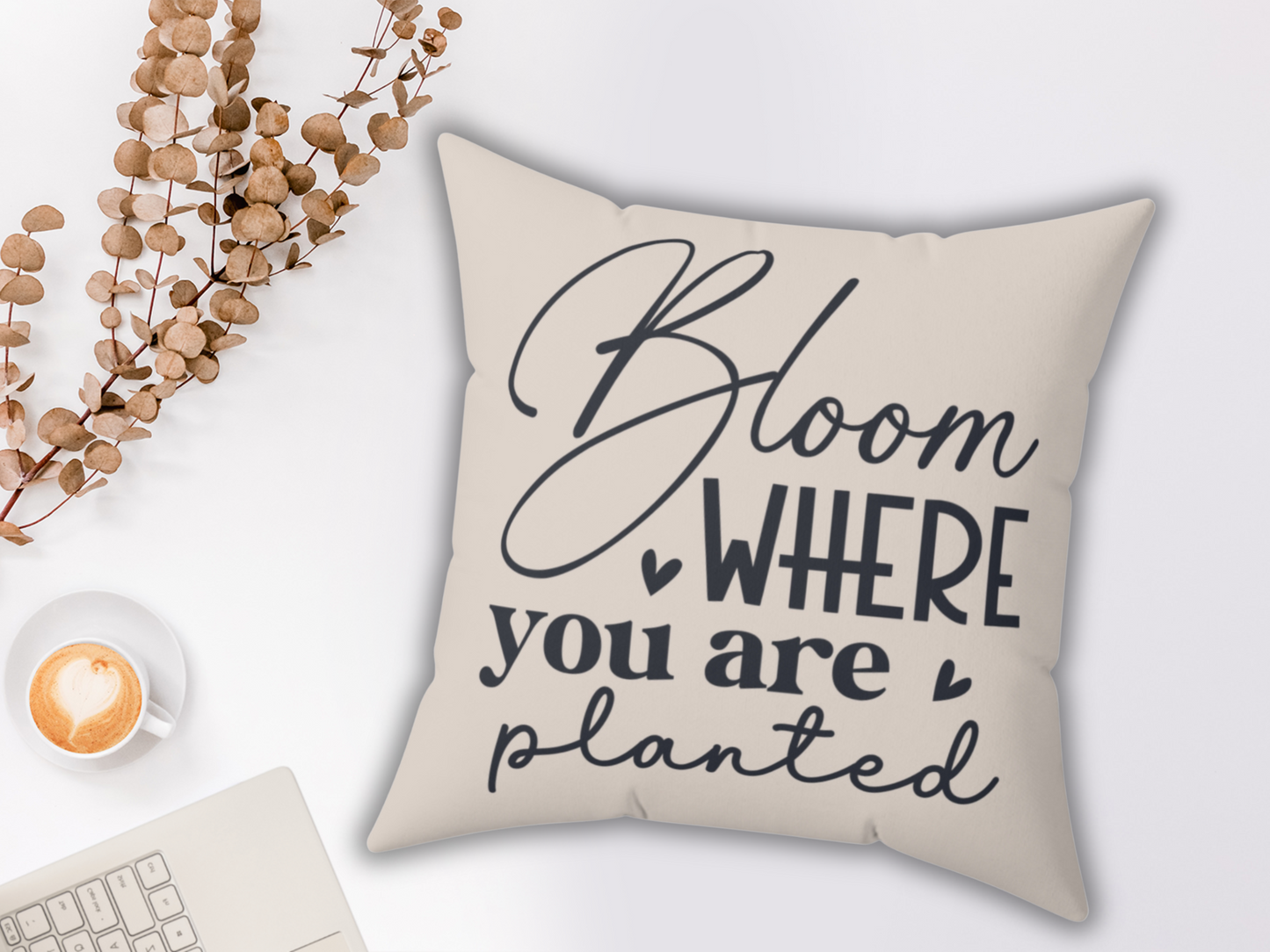 A beige pillow with the text "Bloom where you are planted" in black script. Beside it, dried eucalyptus branches, a latte, and a laptop create a cozy scene.
