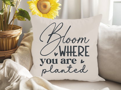 Decorative pillow on a beige sofa with the phrase "Bloom Where You Are Planted" in elegant black script. A vibrant sunflower adds a cheerful touch.
