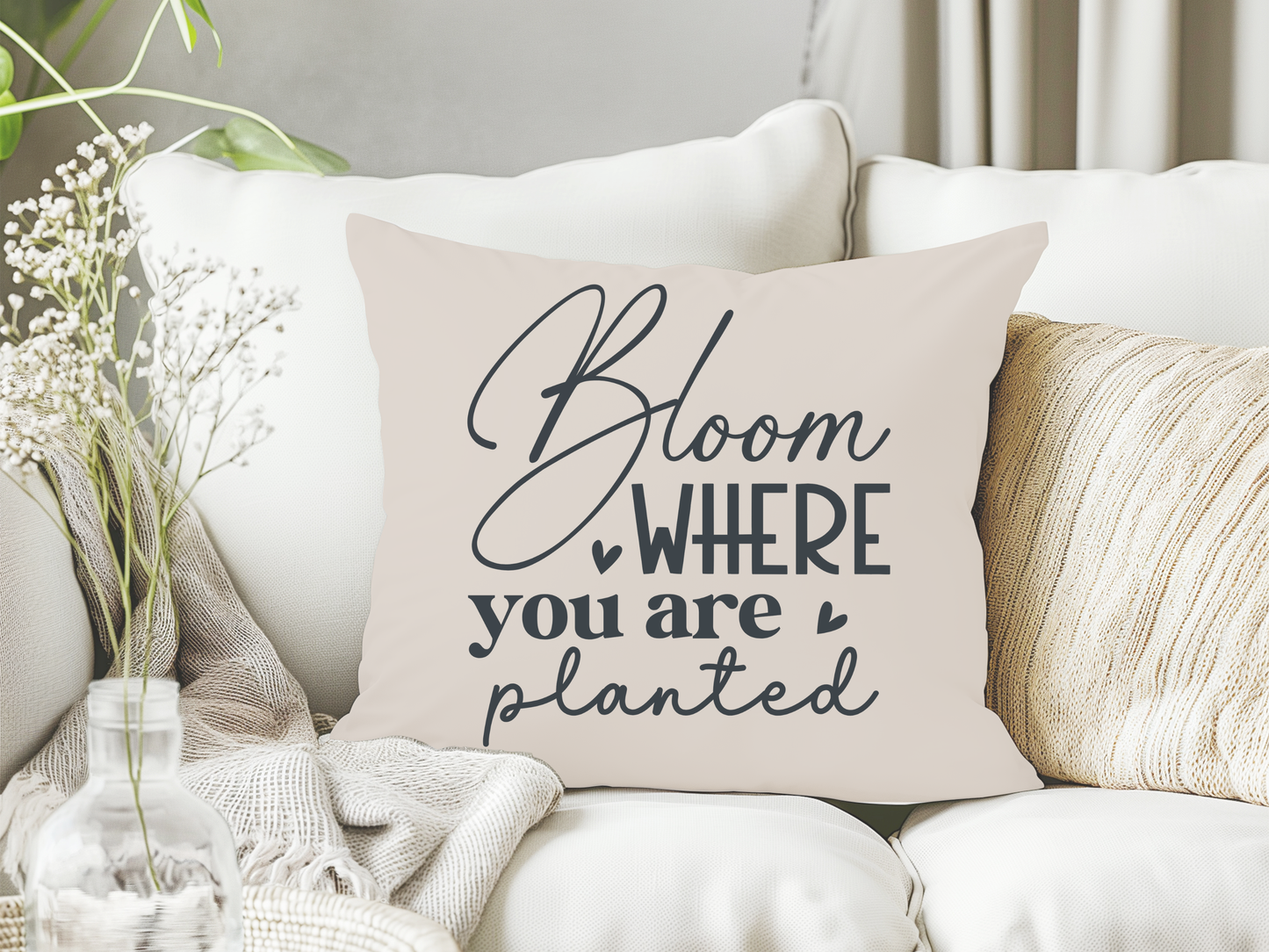 Cushion on a white sofa with "Bloom where you are planted" in elegant script. Nearby, delicate white flowers in a vase, creating a serene vibe.