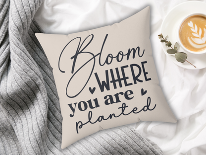 Cream pillow with the text "Bloom where you are planted" in black script. It's placed on a gray knitted blanket. A latte in a white cup sits beside it, creating a cozy atmosphere.