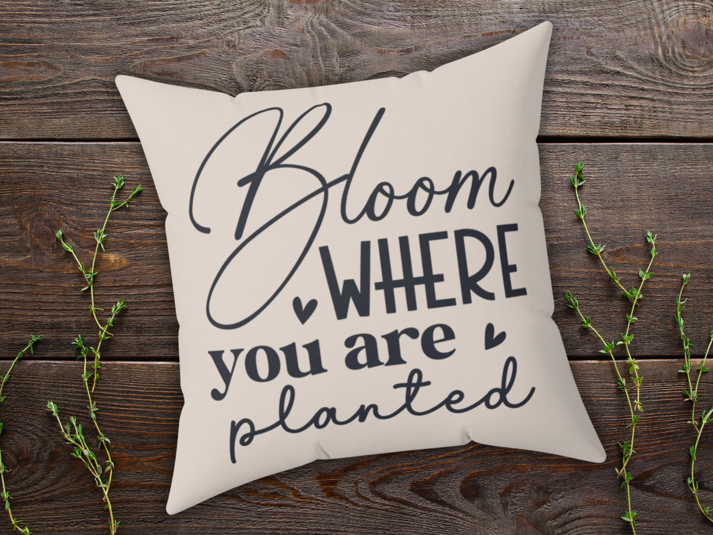 Decorative beige pillow with the text "Bloom where you are planted" in black script. It rests on a rustic wooden surface with sprigs of greenery.