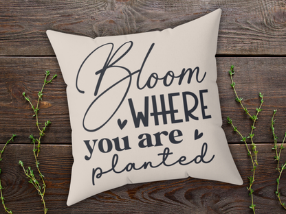Decorative beige pillow with the text "Bloom where you are planted" in black script. It rests on a rustic wooden surface with sprigs of greenery.