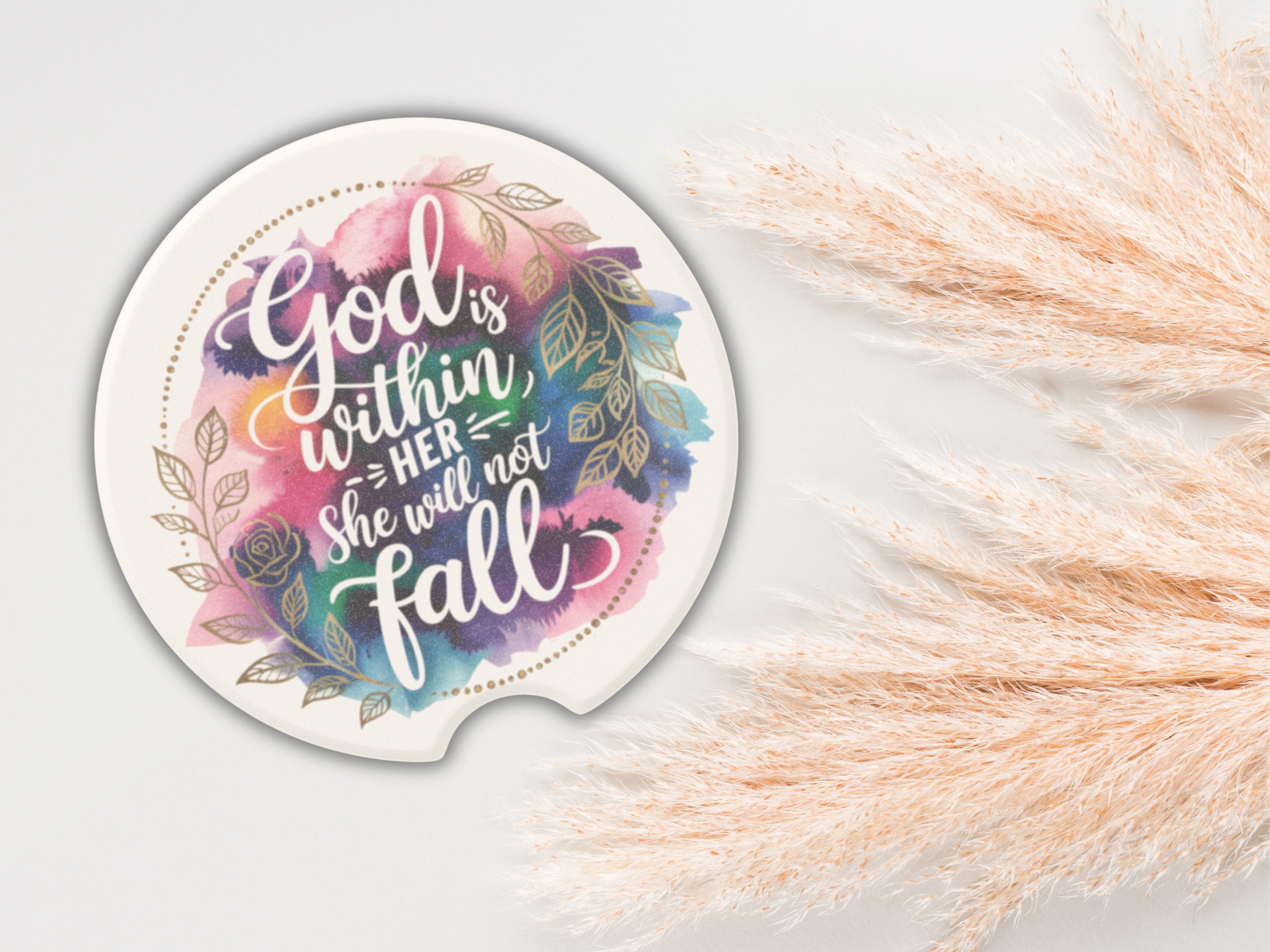 Round coaster with floral design and colorful text: "God is within her, she will not fall." Pampas grass decor on the right adds a serene touch.