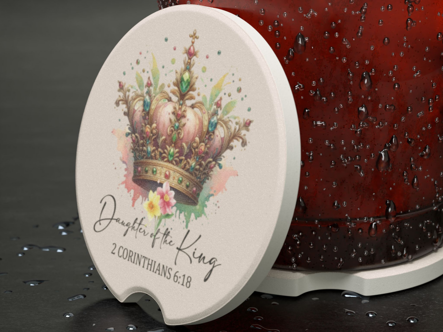 A ceramic coaster with a colorful, jeweled crown illustration and the text "Daughter of the King, 2 Corinthians 6:18," on a wet, dark surface.