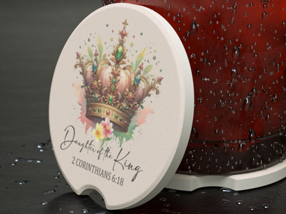 A ceramic coaster with a colorful, jeweled crown illustration and the text "Daughter of the King, 2 Corinthians 6:18," on a wet, dark surface.
