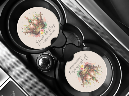 Car cup holder with a decorative coaster featuring a detailed crown illustration, floral accents, and script text reading “Daughter of the King, 2 Corinthians 6:18.”