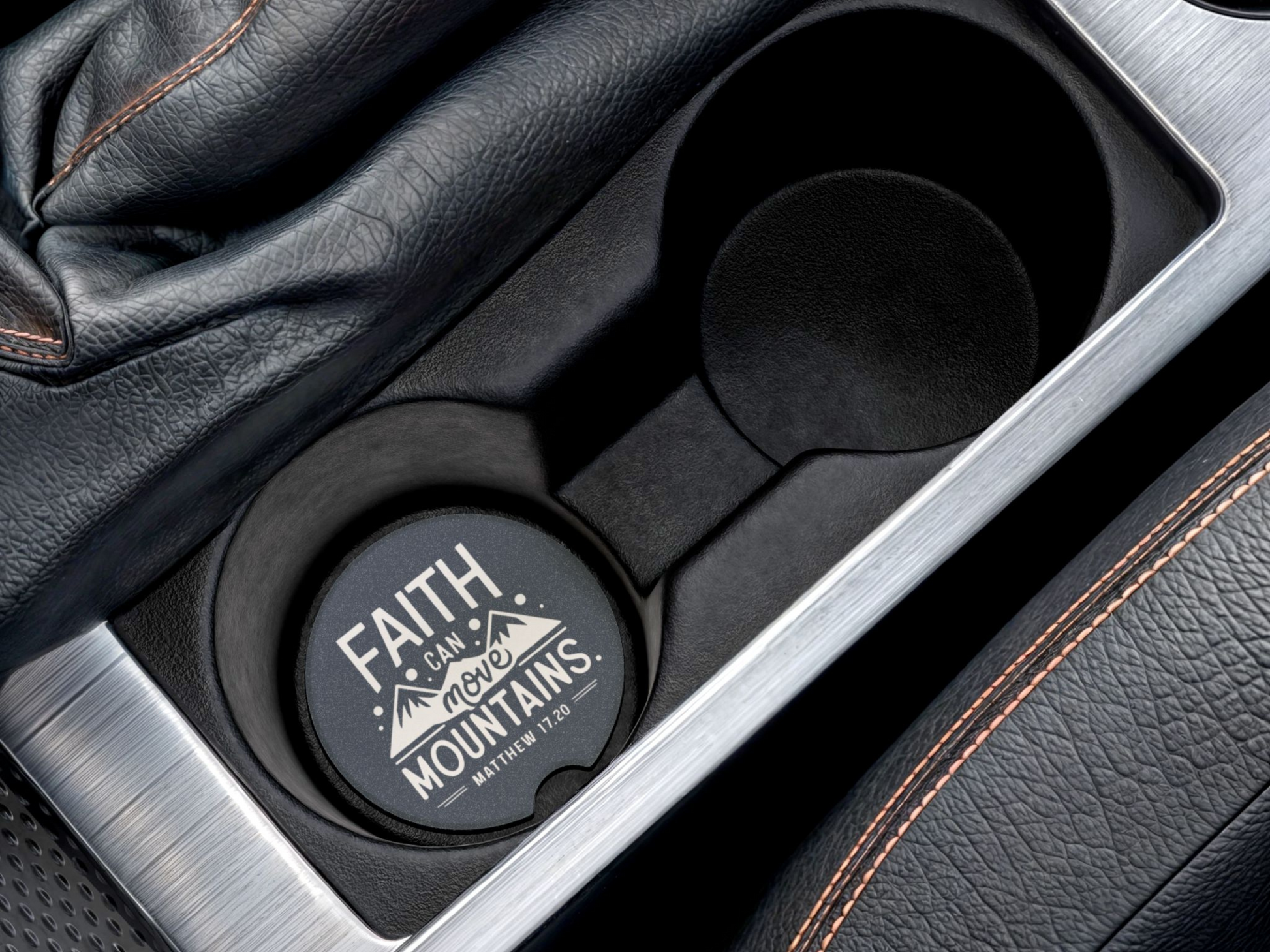 A car cup holder with a coaster that reads "Faith can move mountains," featuring a mountain design. Nearby, a black leather gear shift and seat stitching are visible.