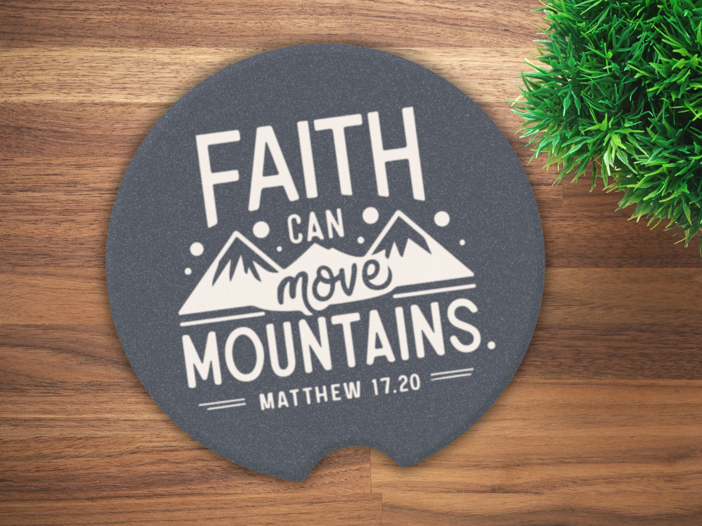 Circular car cup coaster on wooden table with text "Faith can move mountains. Matthew 17:20" alongside mountain illustration. Green plant visible on the side.