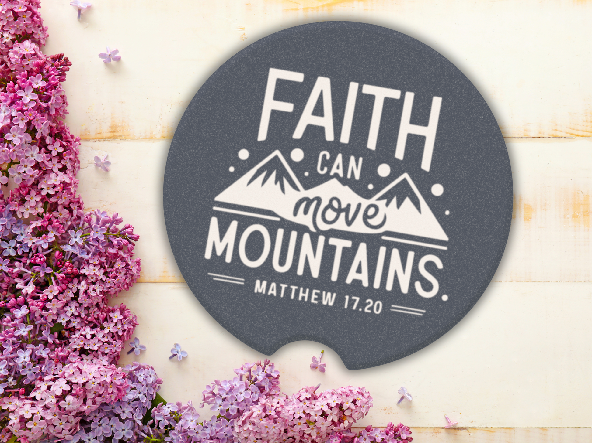 A car cup coaster with the text "Faith Can Move Mountains, Matthew 17:20" in white, featuring mountain graphics. Lilac flowers on the left add a serene touch.