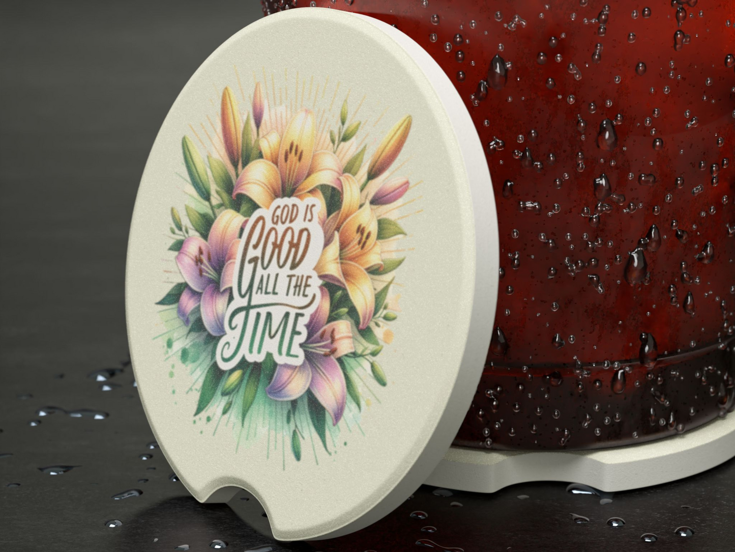 A round coaster with colorful flowers and the text "God is good all the time" is next to a condensation-covered red cup on a wet surface.