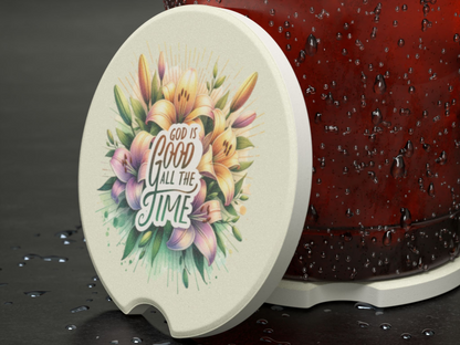 A round coaster with colorful flowers and the text "God is good all the time" is next to a condensation-covered red cup on a wet surface.