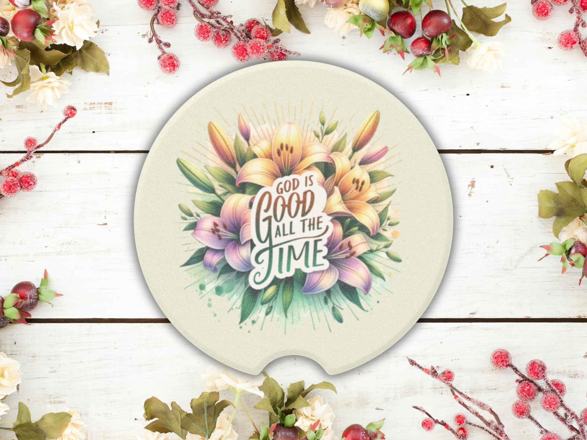 Round Christian car coaster with a floral design featuring lilies, centered on the phrase "God is good all the time." Placed on a wooden surface with decorative berries and foliage.