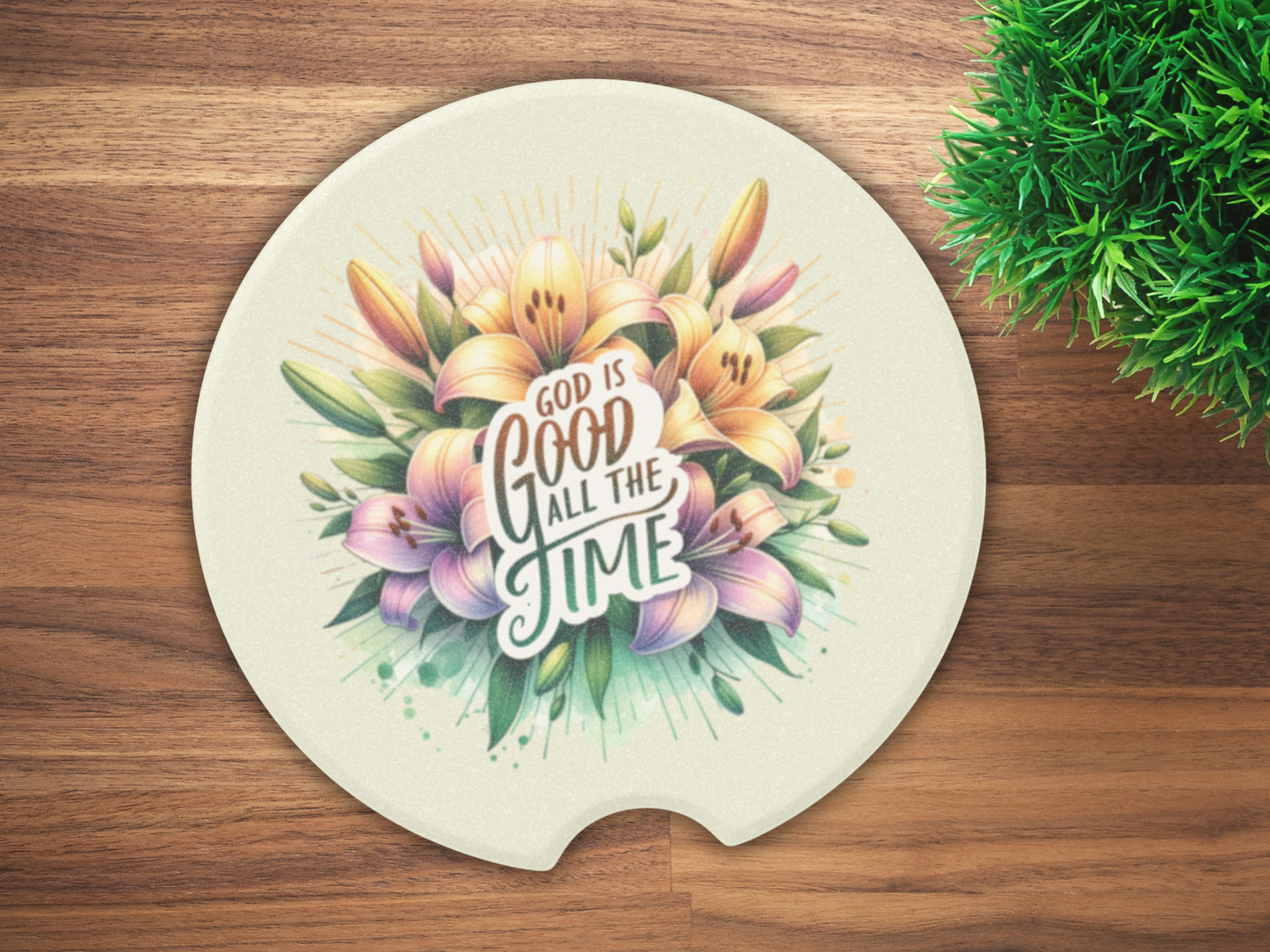 Round car coaster on wooden table, featuring colorful lilies and green leaves. Quote “God is good all the time” is centered. Lush green plant nearby.