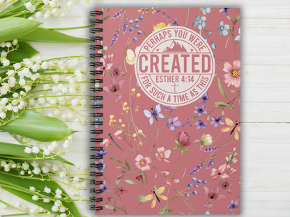 Spiral notebook with floral cover on a wooden surface beside lily of the valley flowers. The cover displays the text, "Perhaps you were created for such a time as this. Esther 4:14." The tone is inspirational and serene.