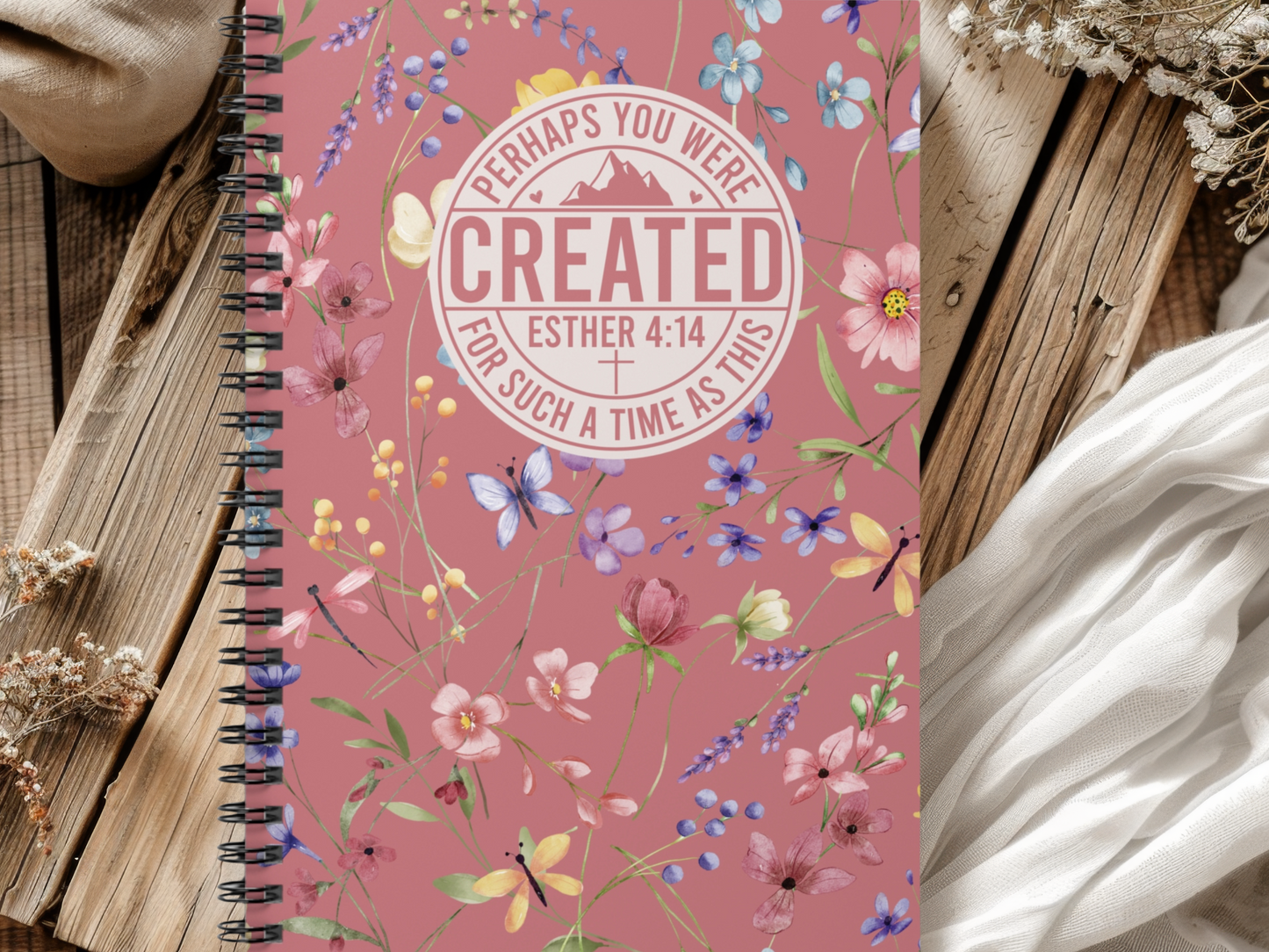 A floral spiral notebook on a wooden table. The pink cover features colorful flowers, butterflies, and a quote: "Perhaps you were created for such a time as this."