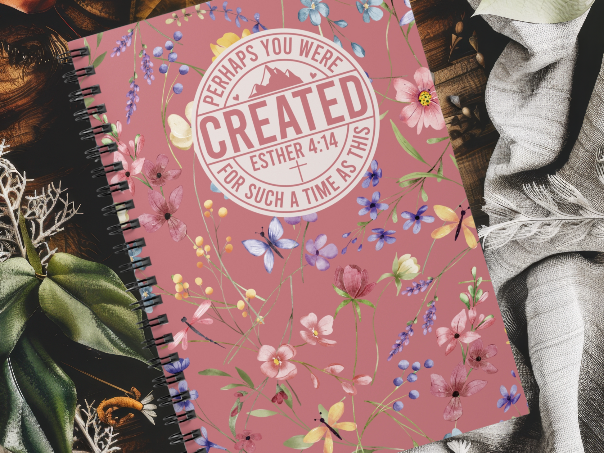 A floral-themed Christian journal for women with a pink cover lies on a table with fabric and greenery. The cover features the text "Perhaps you were created, Esther 4:14, for such a time as this." The design is vibrant and inspirational.