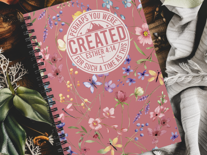 A floral-themed Christian journal for women with a pink cover lies on a table with fabric and greenery. The cover features the text "Perhaps you were created, Esther 4:14, for such a time as this." The design is vibrant and inspirational.