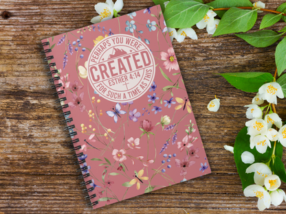 A spiral notebook with the phrase "Perhaps you were created for such a time as this, Esther 4:14" on a rustic wooden table, surrounded by white flowers and green leaves, conveying a serene and motivational tone.
