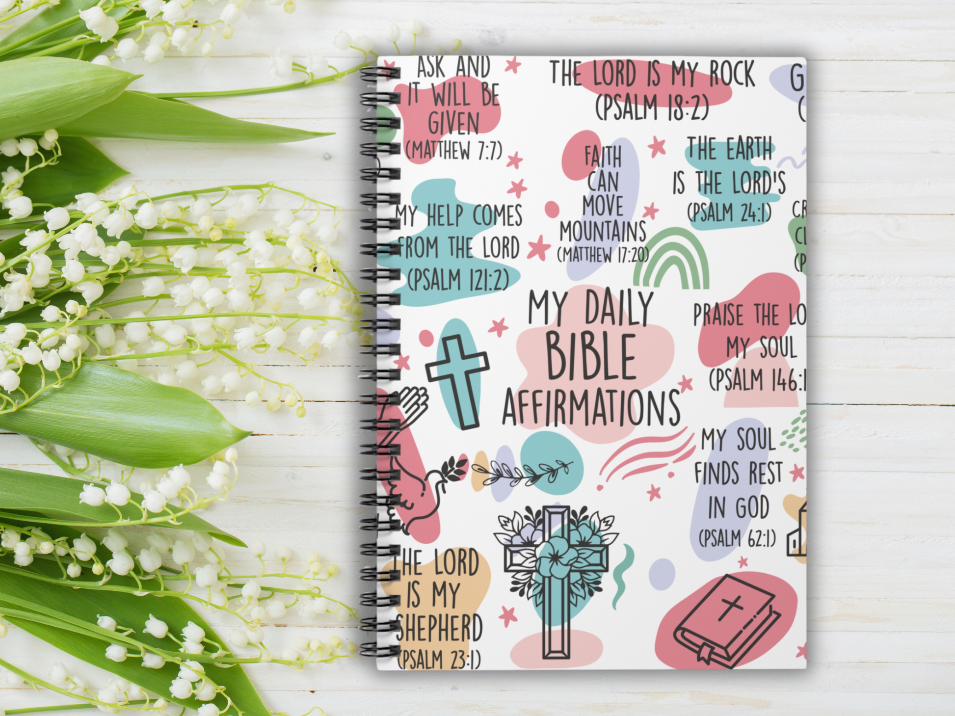 A notebook titled "My Daily Bible Affirmations" with colorful Bible verses on the cover. Surrounding it are delicate white lily of the valley flowers on a white wooden background. The scene conveys peace and inspiration.