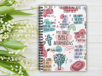 A notebook titled "My Daily Bible Affirmations" with colorful Bible verses on the cover. Surrounding it are delicate white lily of the valley flowers on a white wooden background. The scene conveys peace and inspiration.