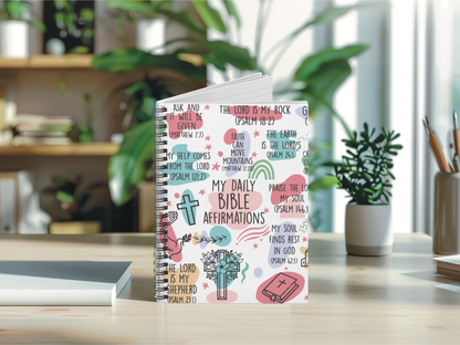 A spiral-bound notebook titled "My Daily Bible Affirmations" on a desk, surrounded by decorative plants and pencils. The cover contains colorful biblical quotes and symbols, creating an uplifting, serene atmosphere.