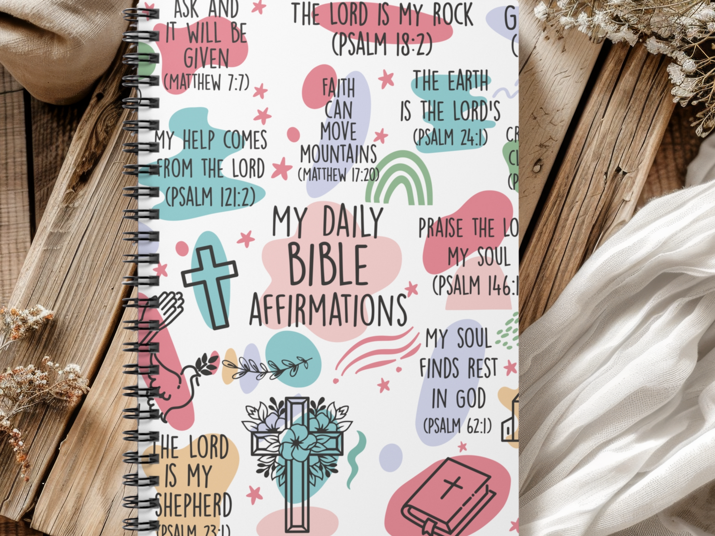 A religious journal on a wooden surface displays colorful, artistic Bible verses and religious symbols like crosses and praying hands, conveying faith and positivity.