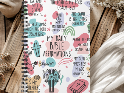 A religious journal on a wooden surface displays colorful, artistic Bible verses and religious symbols like crosses and praying hands, conveying faith and positivity.