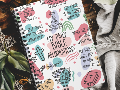 Spiral-bound journal cover with colorful Bible affirmations and symbols like a cross and prayer hands. Lush plants and cozy fabric in the background.