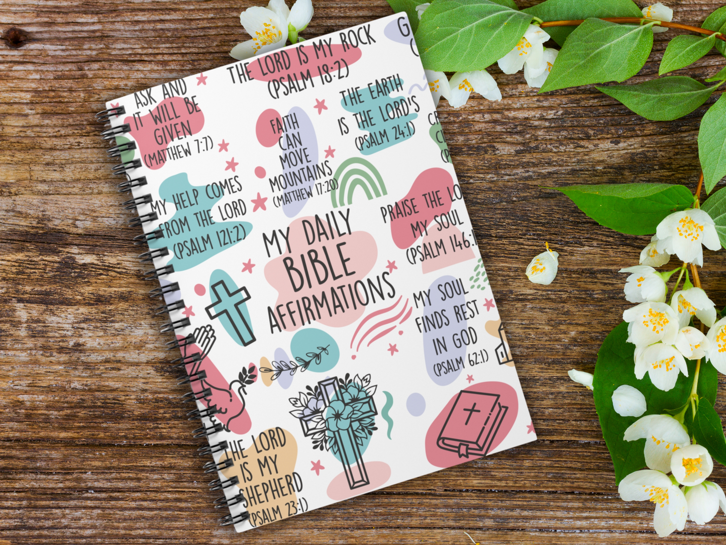 Religious journal on wooden surface, cover with colorful Bible affirmations, icons of crosses, hands, and a Bible. Jasmine flowers on the right for a serene feel.