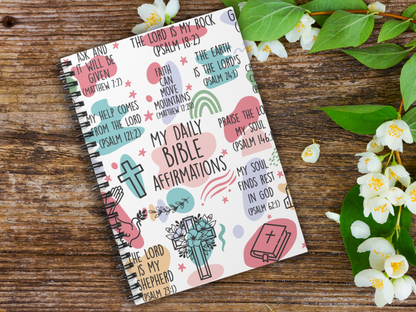 Religious journal on wooden surface, cover with colorful Bible affirmations, icons of crosses, hands, and a Bible. Jasmine flowers on the right for a serene feel.