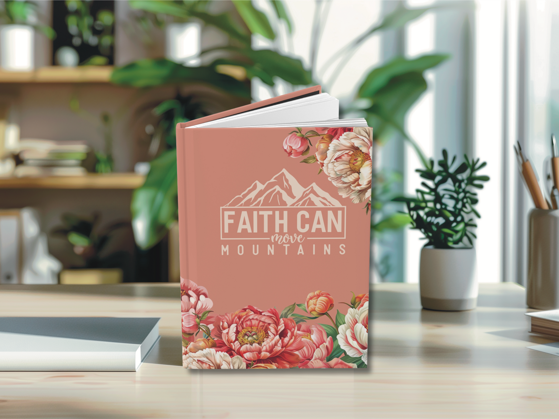 A pink book titled "Faith Can Move Mountains" with floral designs is on a wooden table, surrounded by plants and office supplies, conveying inspiration.