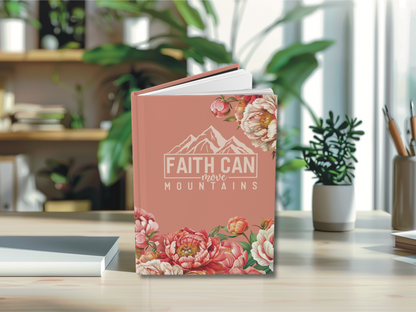 A pink book titled "Faith Can Move Mountains" with floral designs is on a wooden table, surrounded by plants and office supplies, conveying inspiration.
