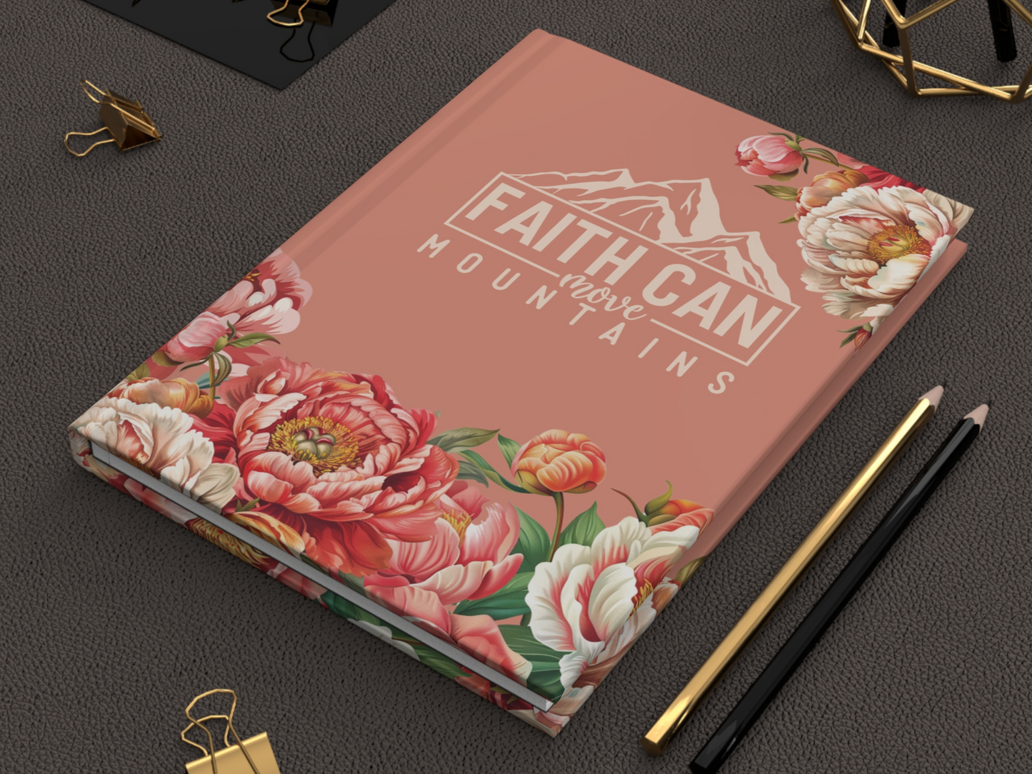 A pink journal with floral designs and the text "Faith Can Move Mountains" lies on a dark surface, accompanied by gold and black pencils and gold paper clips.